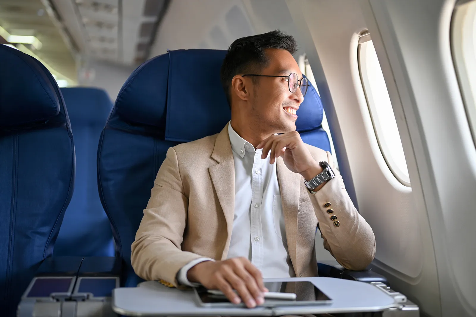 A man wearing a suit sits in a plane with a tablet and pen on the tray table in front of him. He looks out the window, smiling.