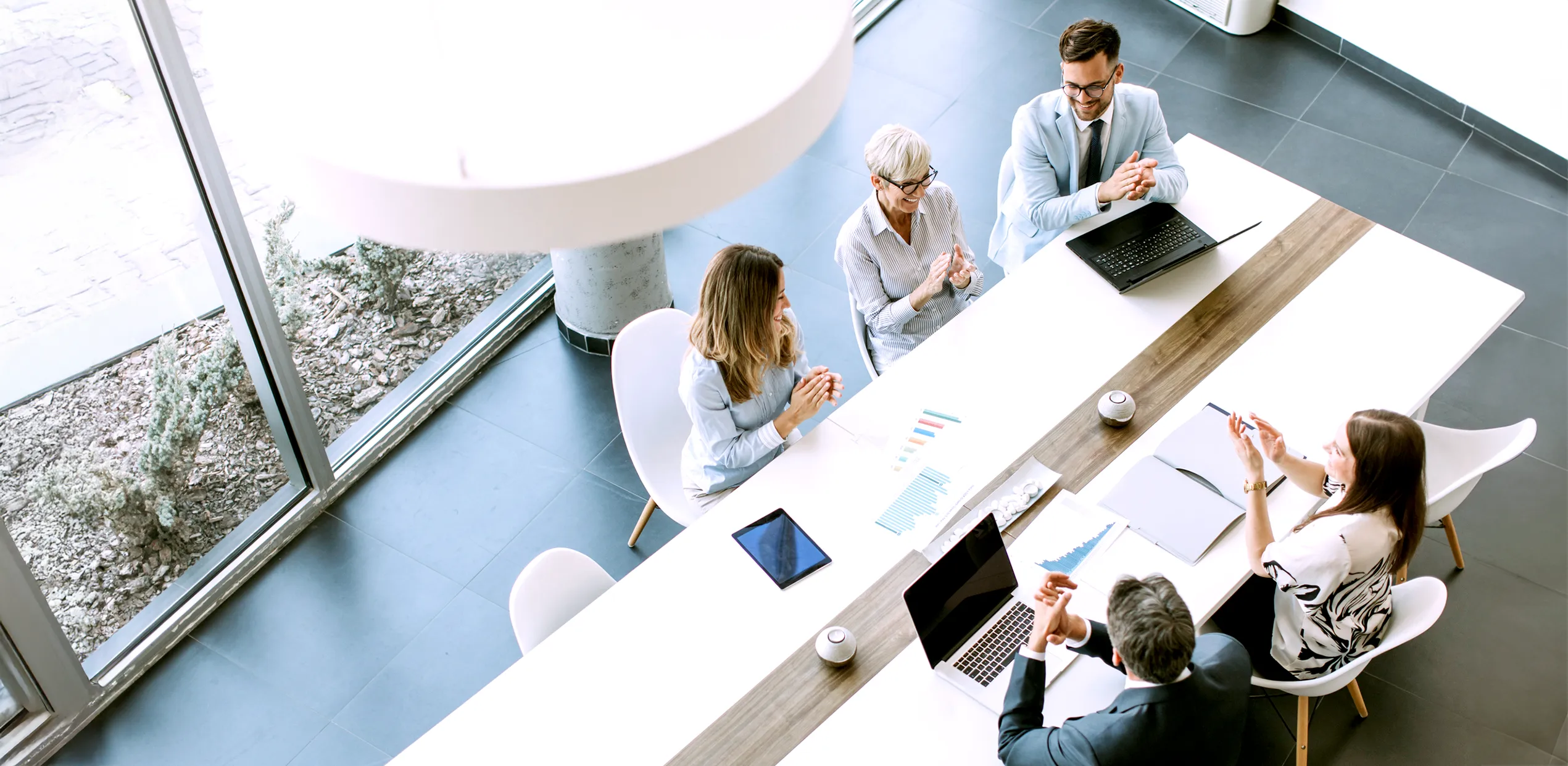 Quatre collègues sont assis autour d'une table de conférence, souriants et applaudissant, avec des ordinateurs et des documents visibles.