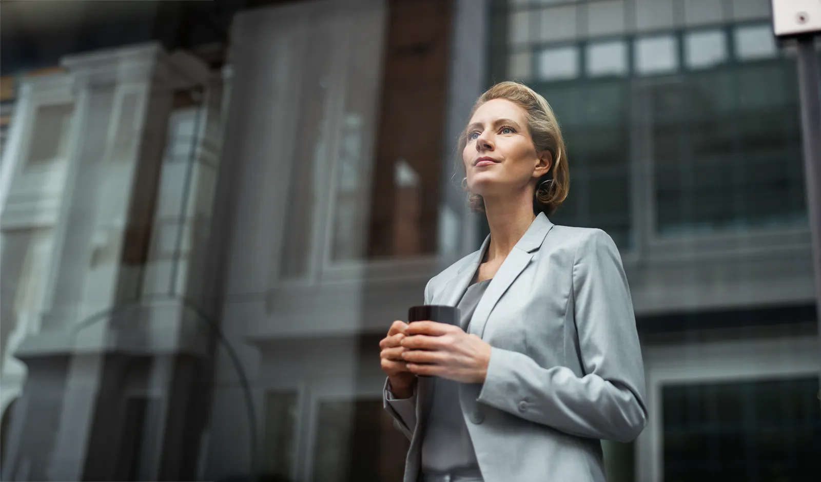 A woman in business attire stands in front of an office building, holding a black porcelain cup.